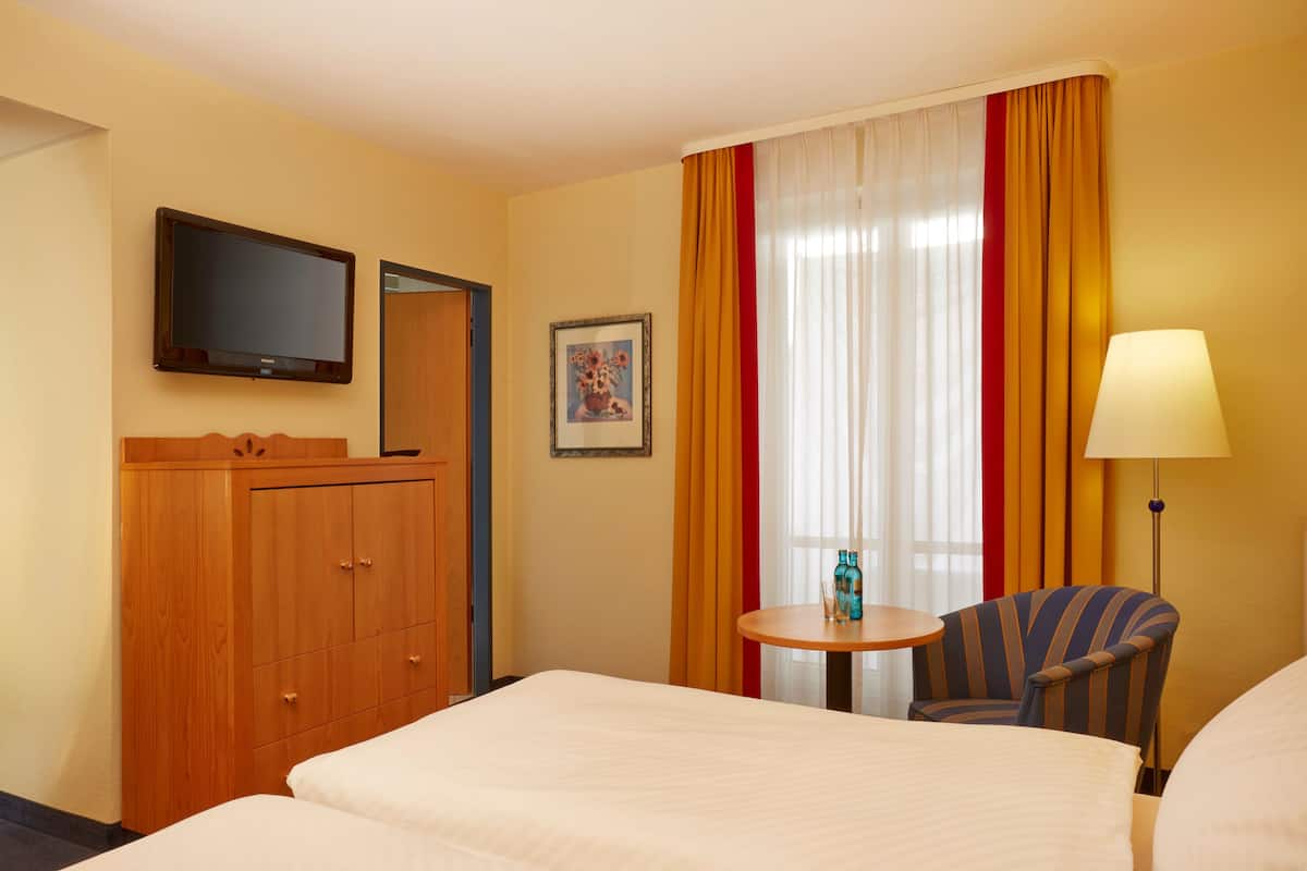 A hotel room with a bed, a TV, a desk with a chair, and a window with curtains.