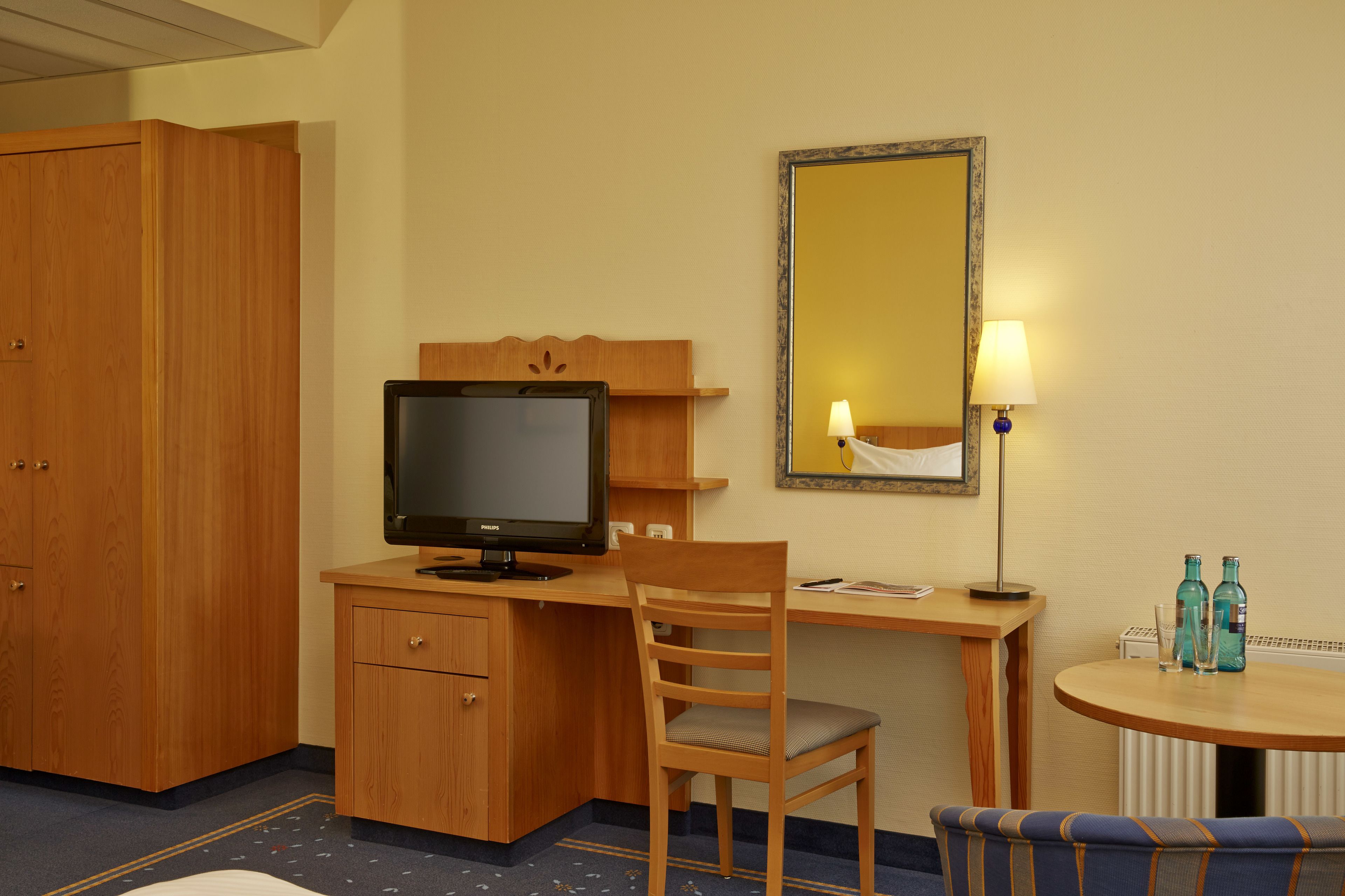 Une chambre d’hôtel avec un bureau en bois, une télévision, une chaise, une table ronde avec deux bouteilles et un miroir au mur.