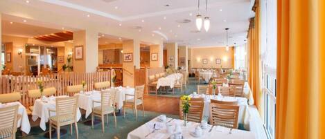 A spacious dining area with multiple tables and chairs, a high ceiling with recessed lighting, and a view of the dining area through a glass partition.