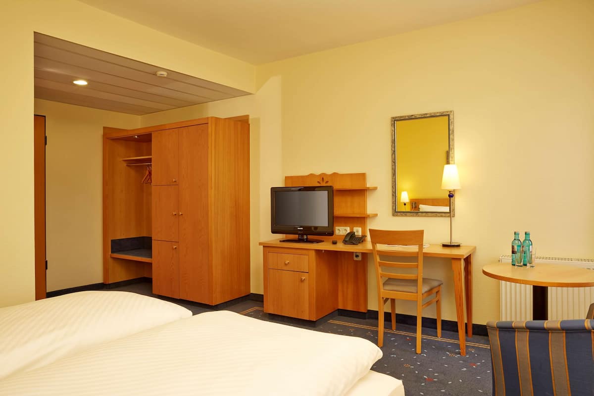 A hotel room with a bed, a desk with a television, a chair, a small table with bottles, and a mirror on the wall.