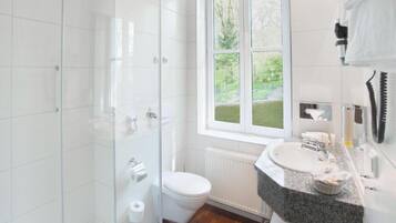 A modern bathroom with a glass shower enclosure, a toilet, a sink with a mirror, and a window with a view of greenery.