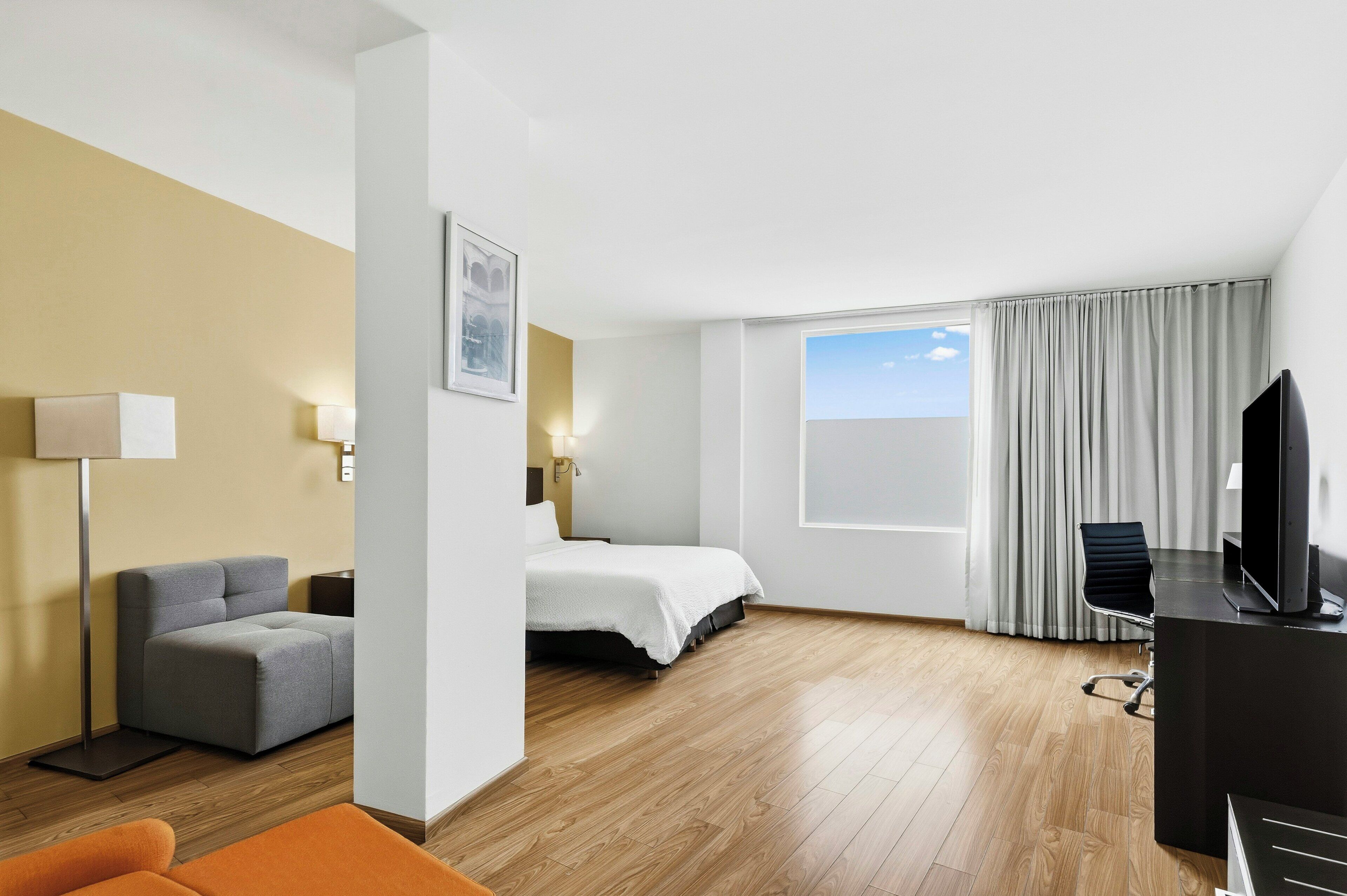 A hotel room with a bed, a desk with a TV, a chair, a sofa, and a window with curtains.