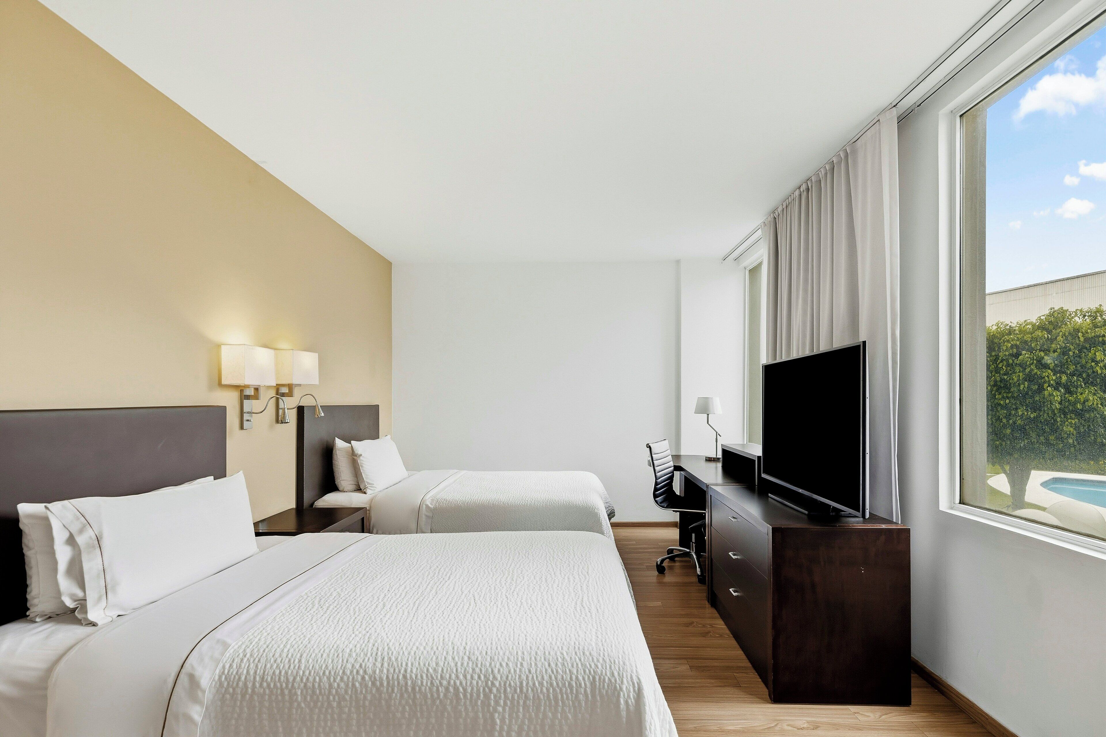 A hotel room with two beds, a desk with a TV, and a window with a view of greenery.