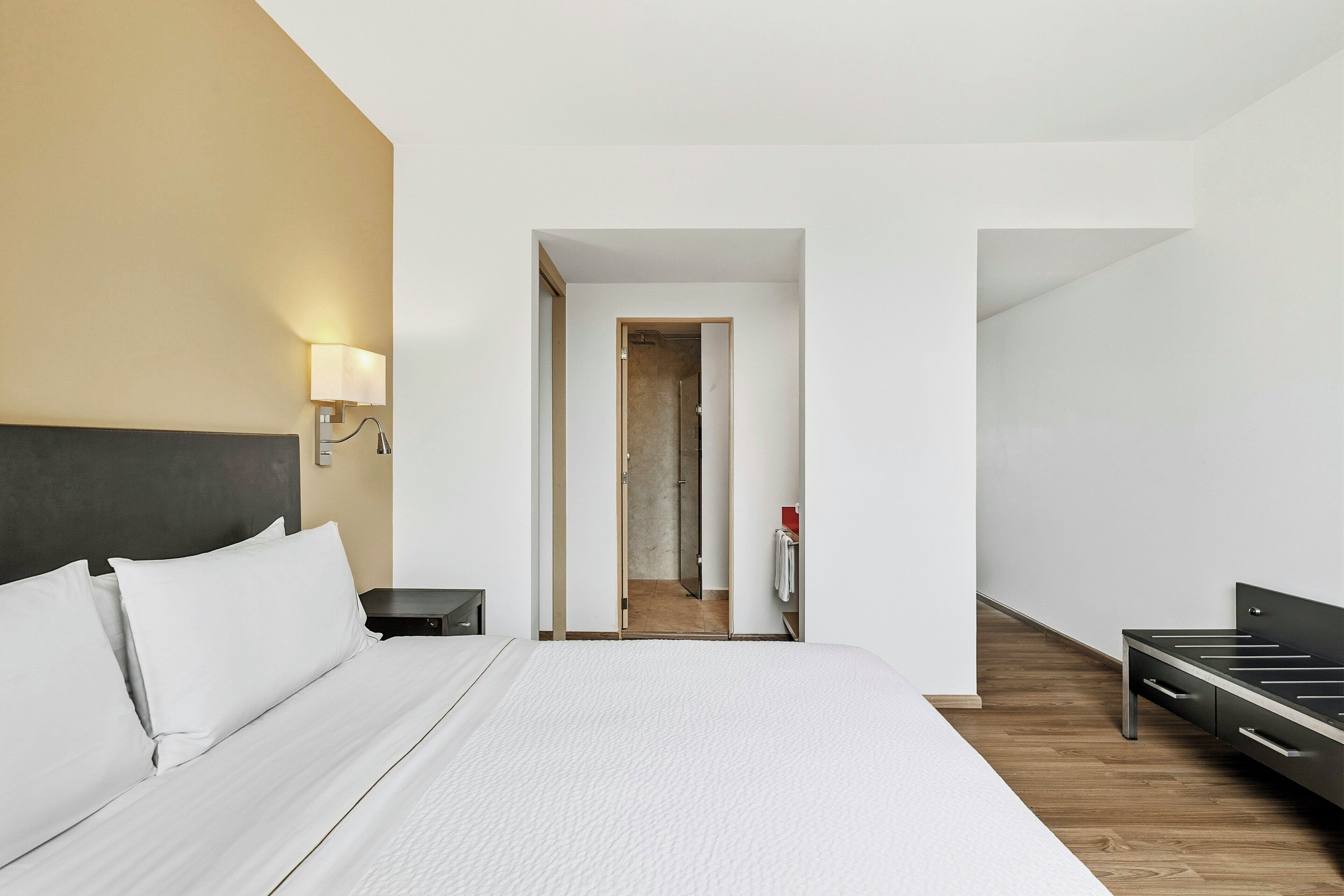 A hotel room with a bed, bedside table, lamp, and a view into the bathroom.
