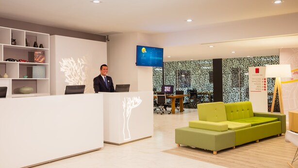 Zona d'entrada d'un hotel modern, amb un taulell de recepció blanc, una pantalla digital que mostra el temps i un sofà verd.