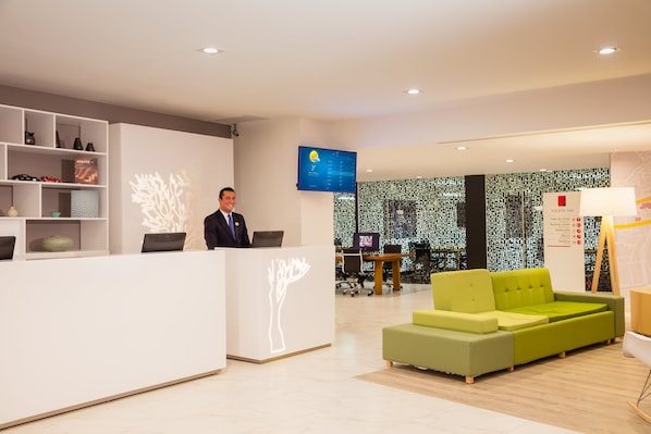 A modern hotel reception area with a white reception desk, a digital screen displaying the weather, and a green sofa.
