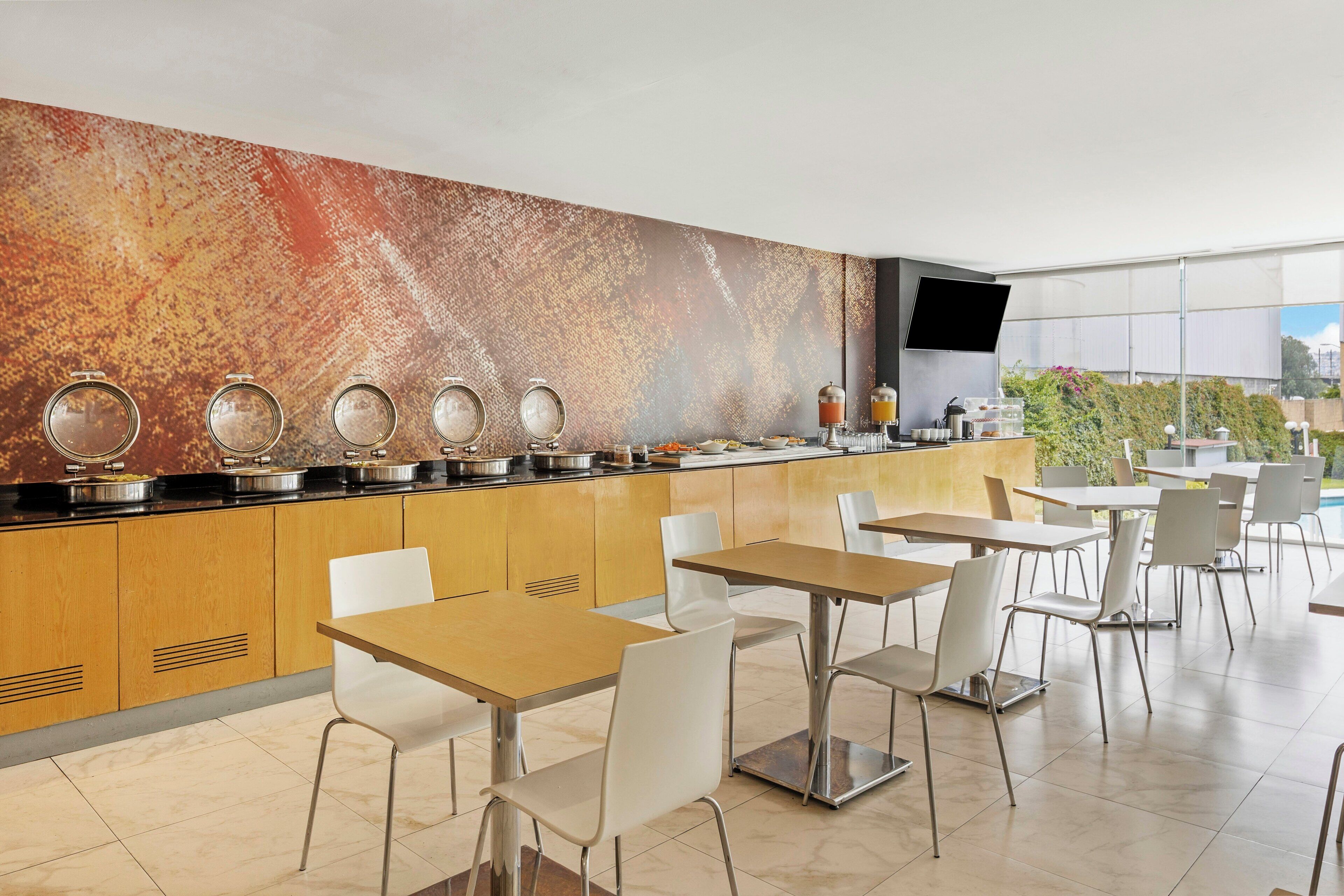 A modern kitchen with a buffet setup, white chairs, and a large window.