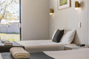 A hotel room with a bed, a bedside table, a wall-mounted light, and a window with a view of greenery outside.