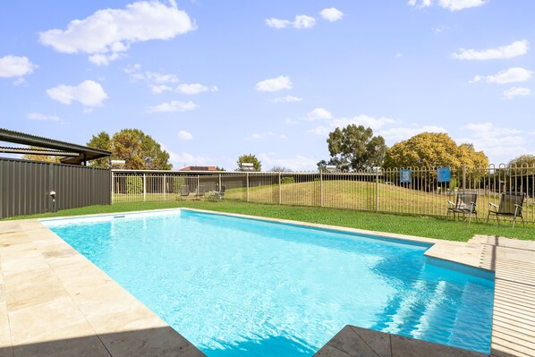 A rectangular swimming pool with a tiled deck, surrounded by a fence and a grassy area with trees.