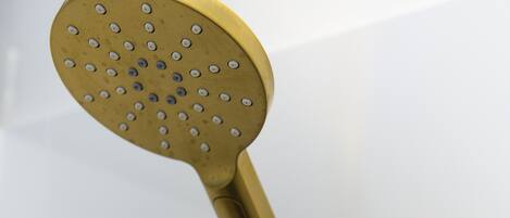 Un cabezal de ducha dorado de diseño redondo y con múltiples boquillas, unido a una tubería vertical mediante una manguera flexible.