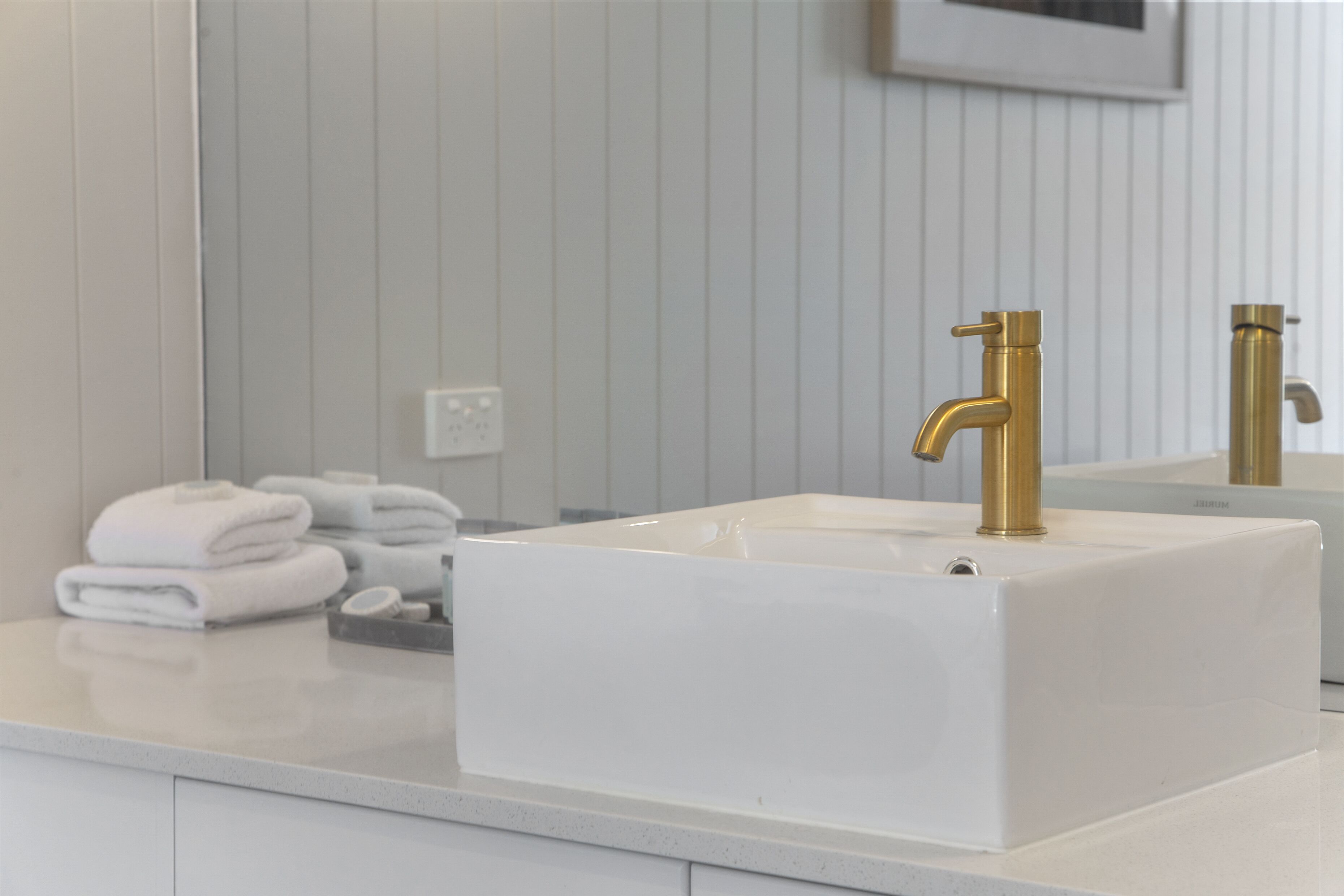 Ein Badezimmer mit einem weißen Waschbecken, goldenen Wasserhähnen und einem Spiegel, der den Raum widerspiegelt.