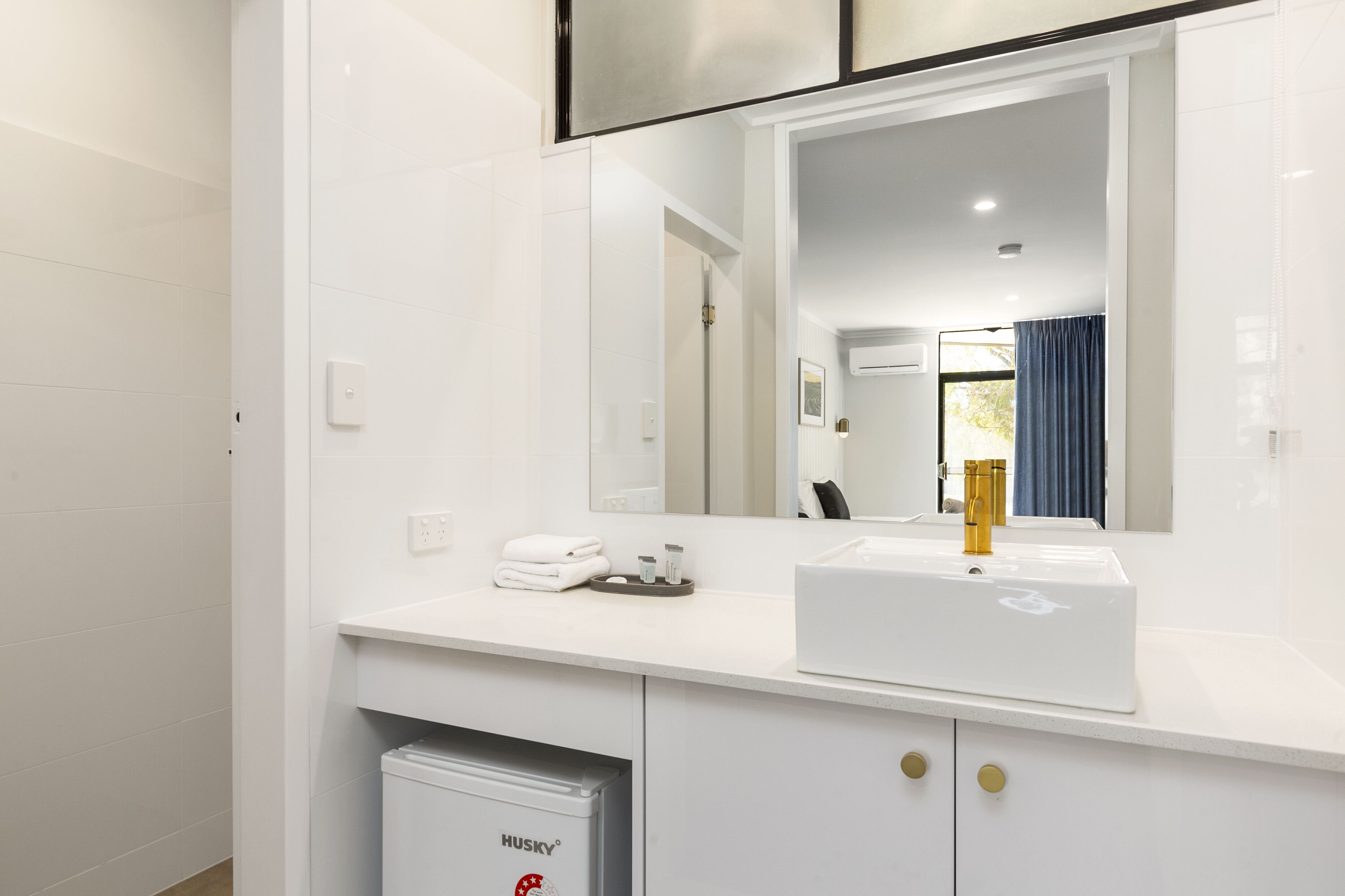Casa de banho moderna com lavatório branco, torneira dourada e espelho em cima. Há um porta-toalhas com toalhas dobradas e um pequeno contentor de lixo com a etiqueta "HUSKY" abaixo do lavatório.