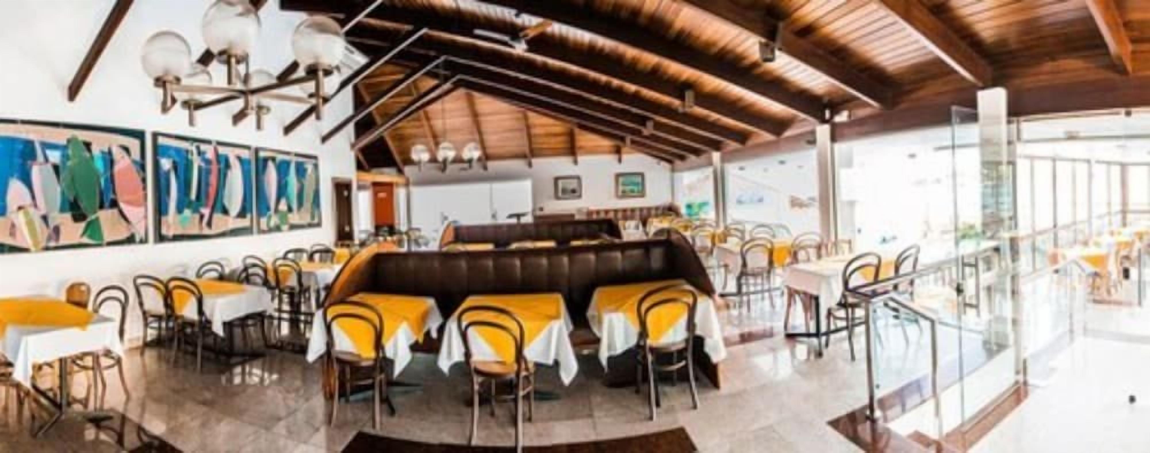 Restaurante com teto alto, vigas de madeira e área de estar central com mesas e cadeiras.