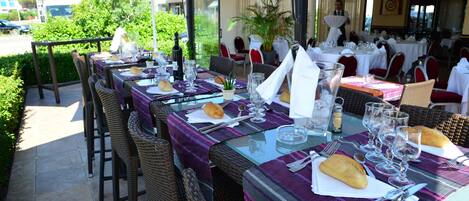 An outdoor dining area with tables set for two, featuring wicker chairs and purple tablecloths.