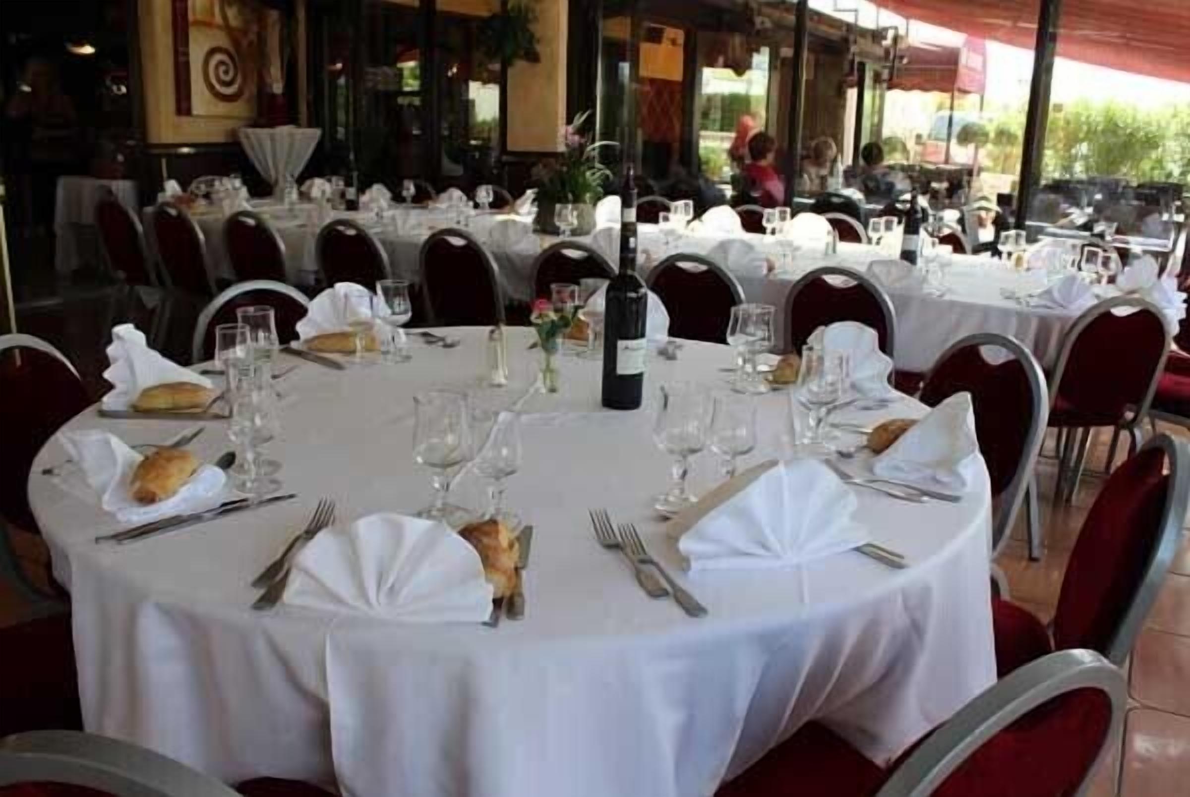 A restaurant with round tables set for dining, each with white tablecloths, red chairs, and neatly arranged cutlery and glasses.