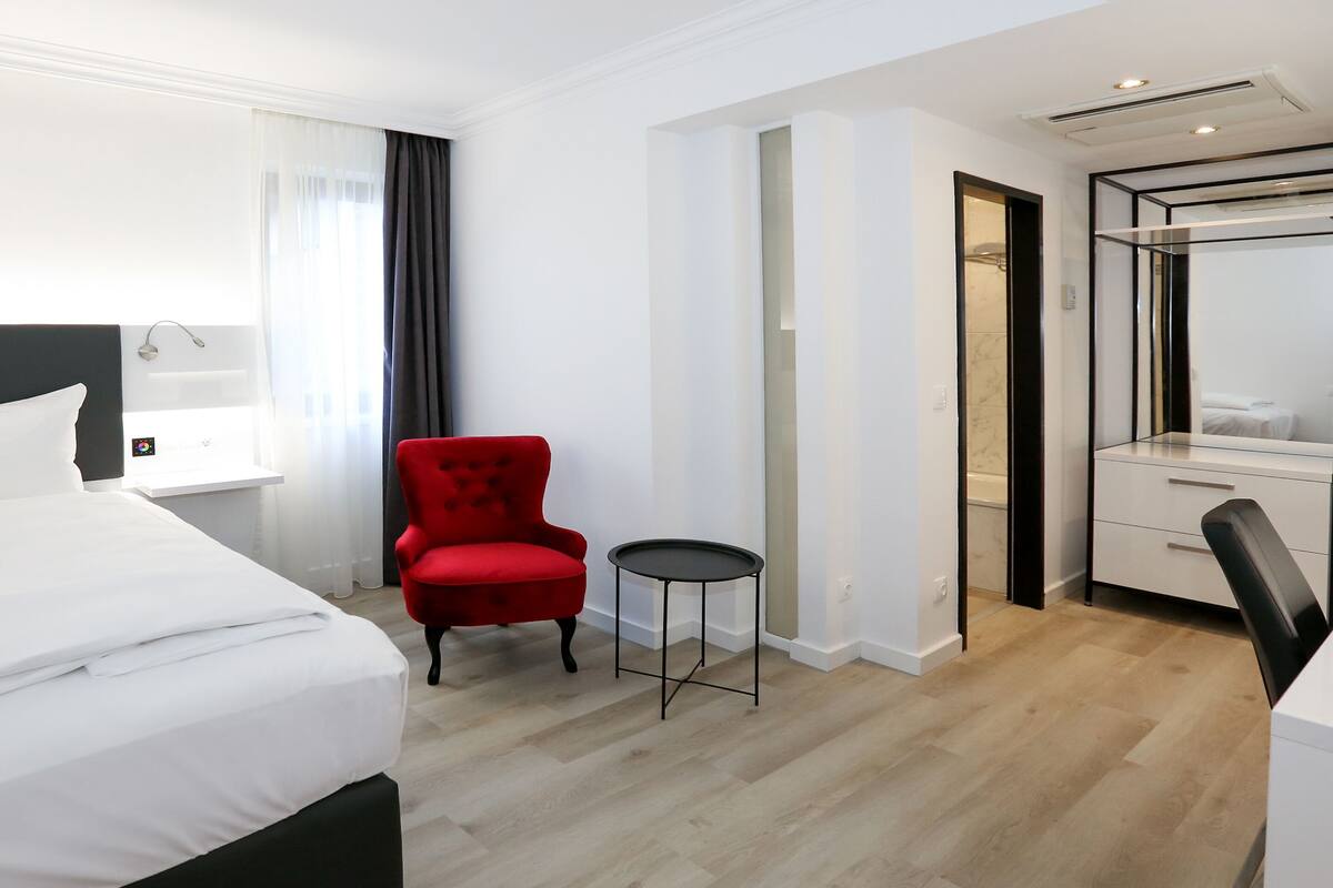 A modern hotel room with a large bed, a red armchair, a small round table, a desk with a mirror, and a walk-in closet.