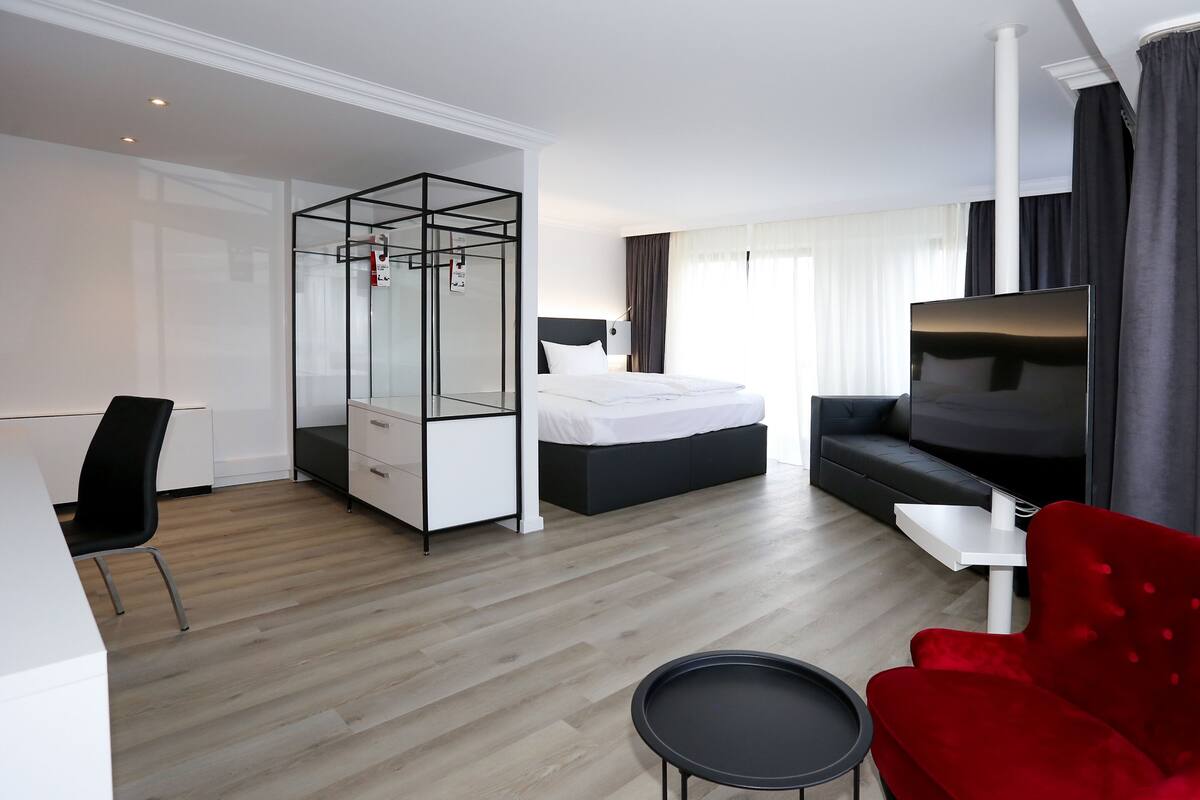 A modern hotel room with a bed, a TV, a red chair, a small table, and a walk-in closet.