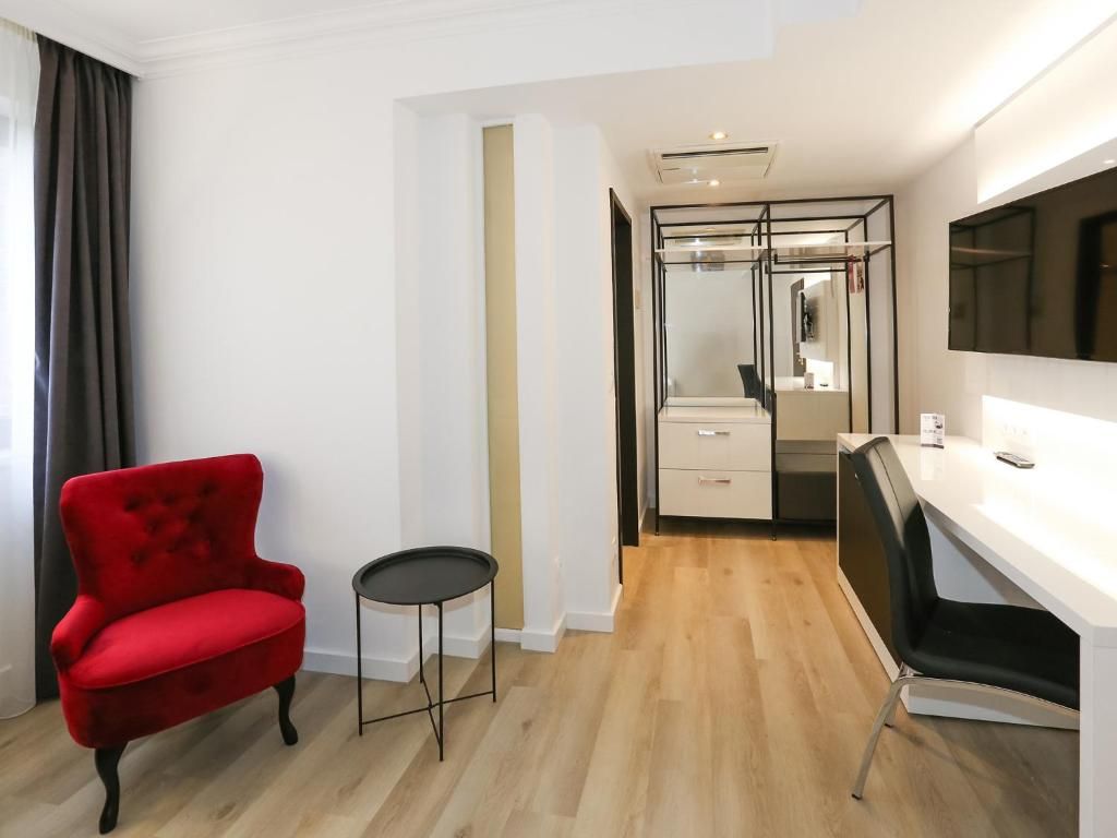 Una habitación de hotel moderna con un sillón rojo, una mesita redonda pequeña, un escritorio blanco y un espejo.
