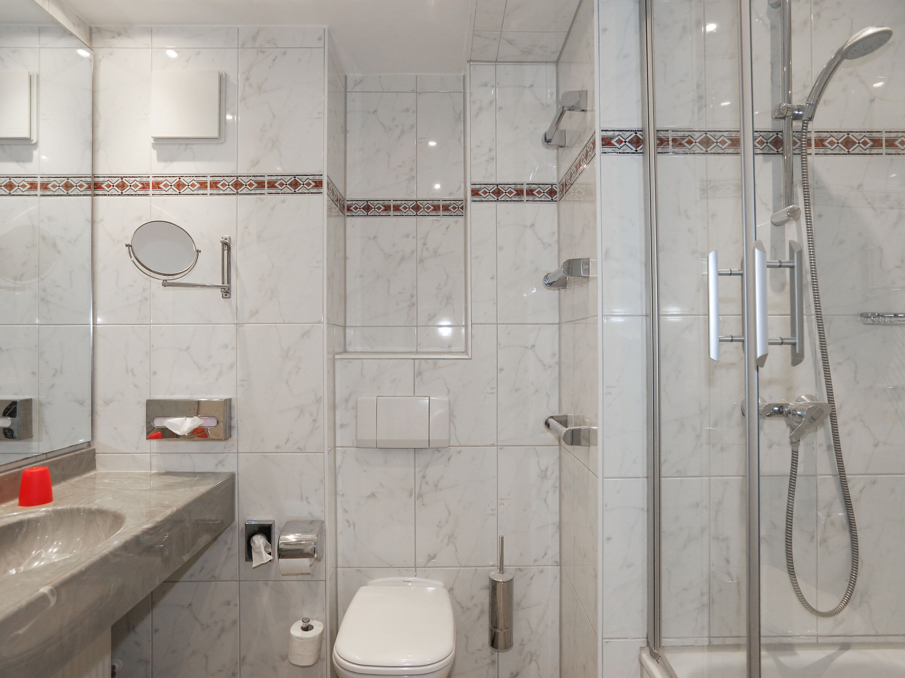A modern bathroom with a glass shower enclosure, a marble-tiled wall, a sink with a red cup, and a toilet.