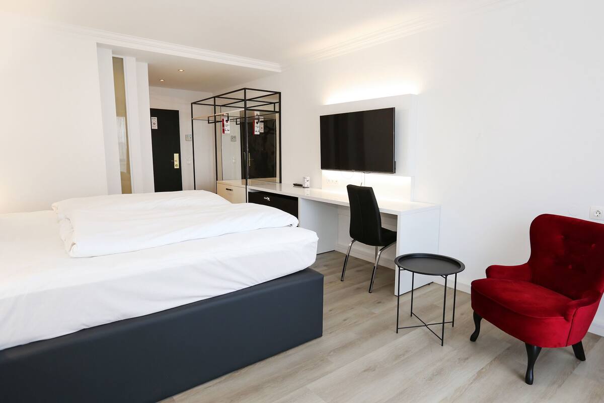 A modern hotel room with a bed, a desk, a chair, and a red armchair.