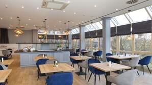 A modern restaurant with a central island, blue and grey chairs, wooden tables, and large windows allowing natural light. - DORMERO Hotel Meerbusch (Meerbusch)