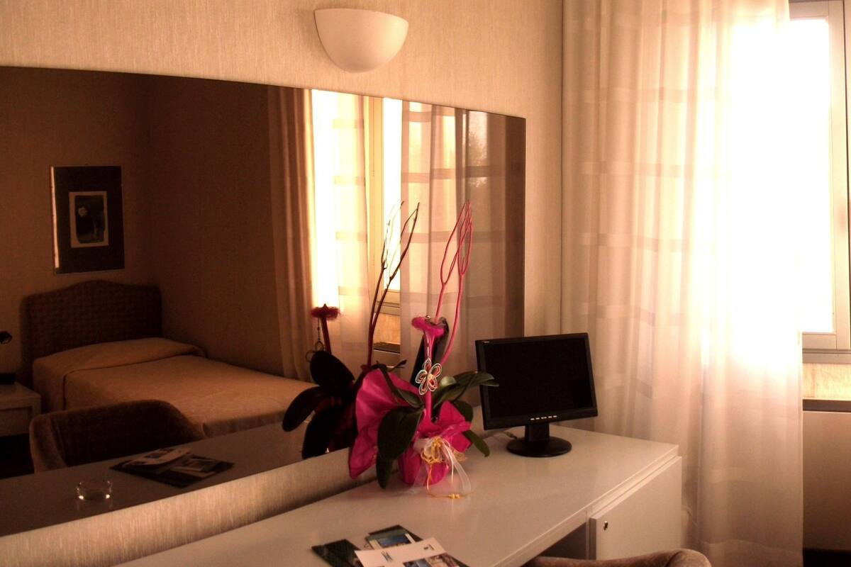 Une chambre d’hôtel avec un lit, un bureau avec un ordinateur, un miroir et une fenêtre avec des rideaux.