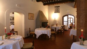 Un comedor con mesas redondas, manteles blancos y velas. Hay sillas alrededor de las mesas, y se ve una chimenea al fondo.