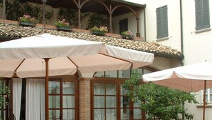 Un patio al aire libre con mobiliario de mimbre y sombrillas blancas, una planta en maceta y un edificio con techo de tejas y ventanas.