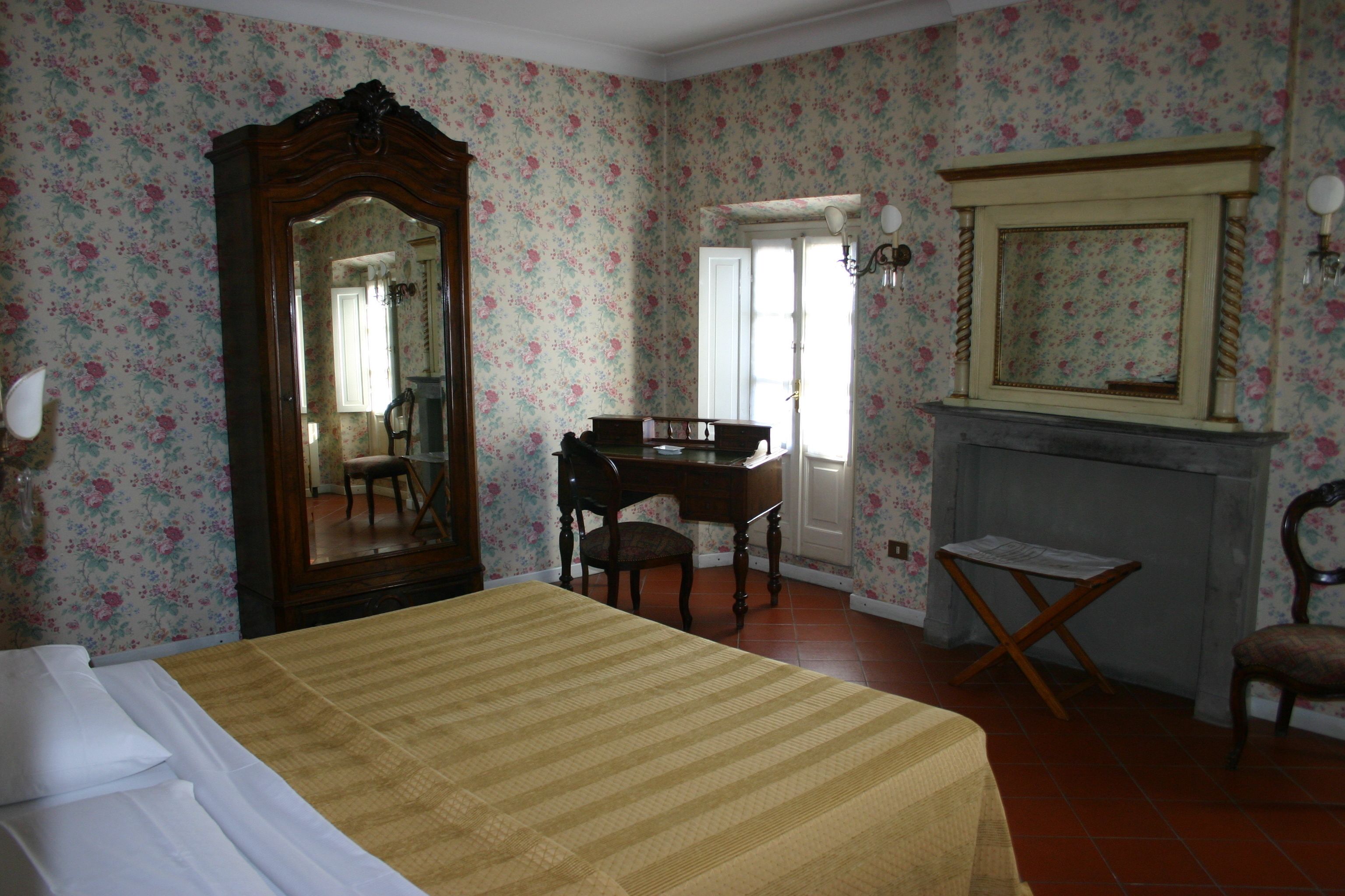 Ein Schlafzimmer mit floraler Tapete, einem großen Bett, einem Holztisch, einem Sessel, einem Kamin und einem Spiegel.
