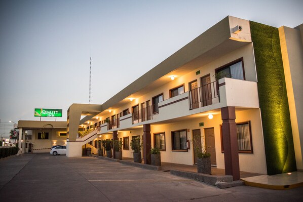 Un hotel de varios pisos con un letrero verde, iluminación exterior y un estacionamiento.