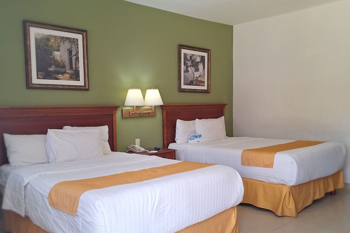 A hotel room with two beds, green walls, framed pictures, and a nightstand with a lamp.