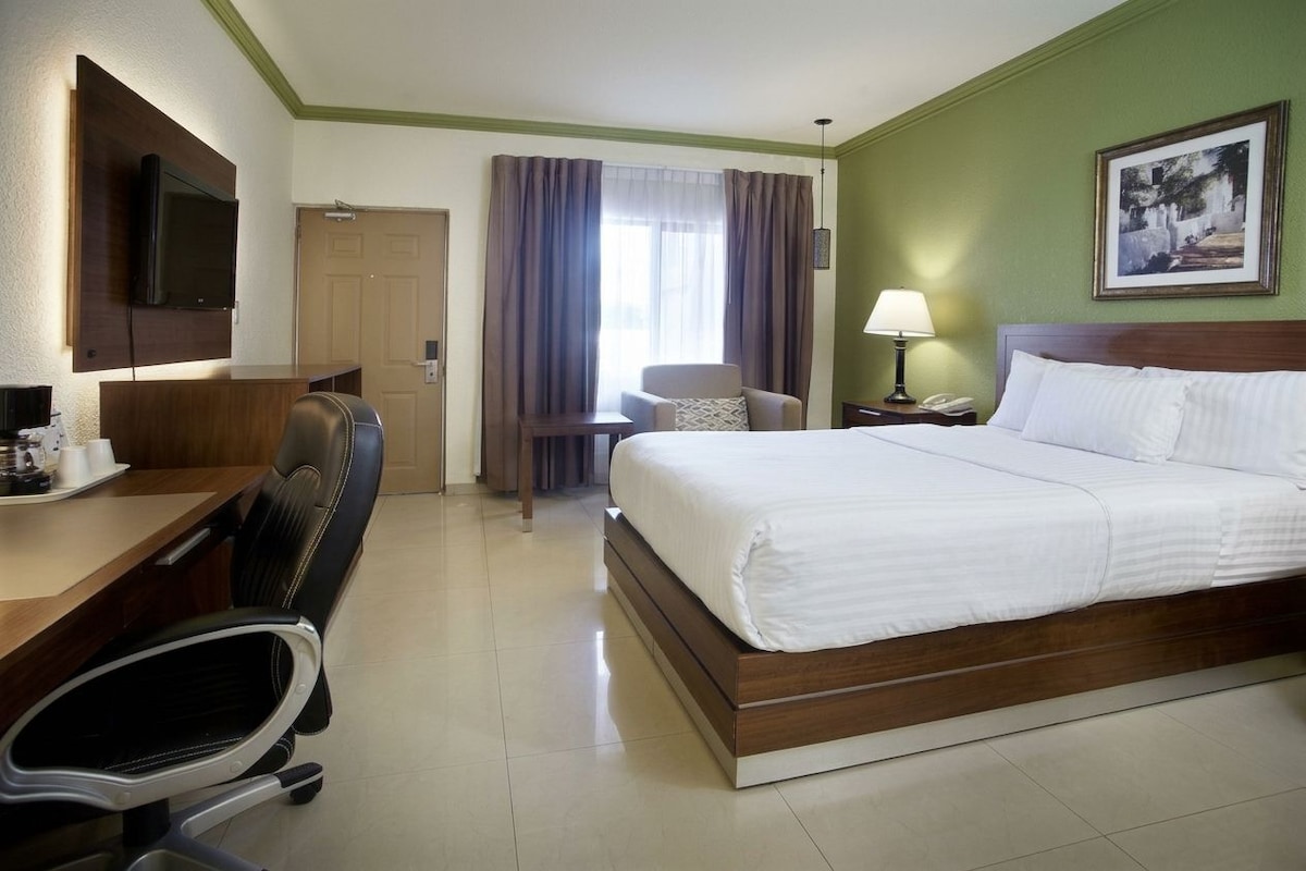 Habitación de hotel con cama, escritorio, silla, televisión y ventana con cortinas.