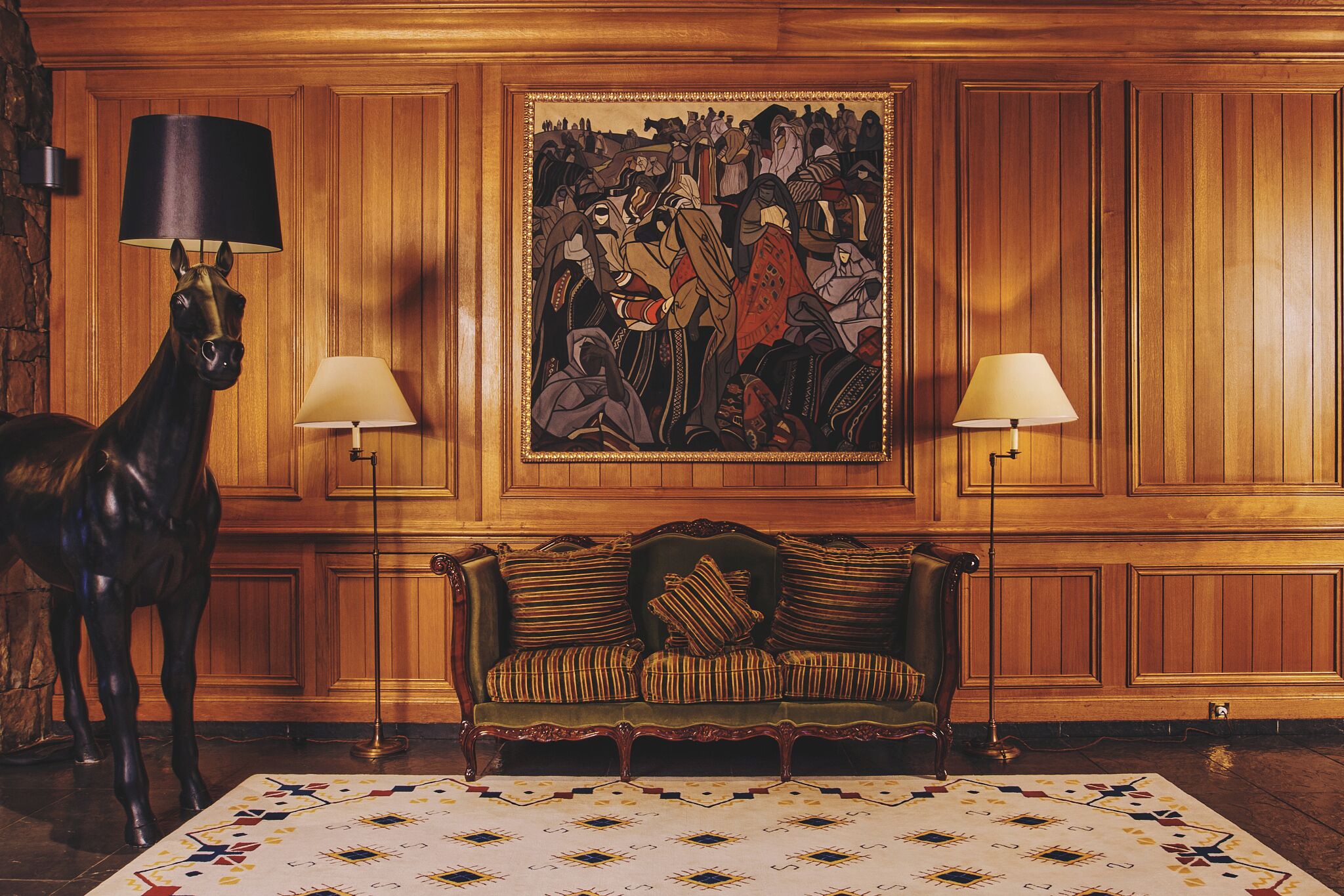 A room with a wooden paneled wall, a green upholstered sofa, two standing lamps, and a horse statue.