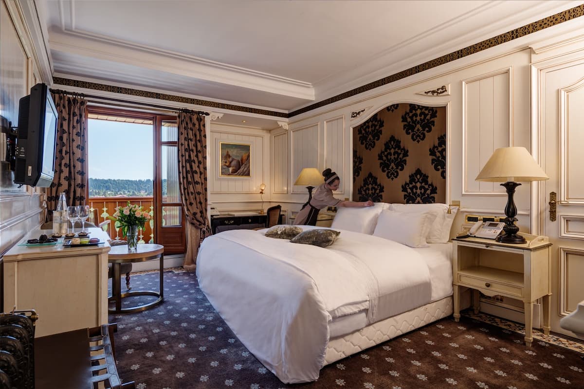 A hotel room with a large bed, a desk with a computer, a chair, a table with a vase of flowers, a lamp, and a view of the outdoors.