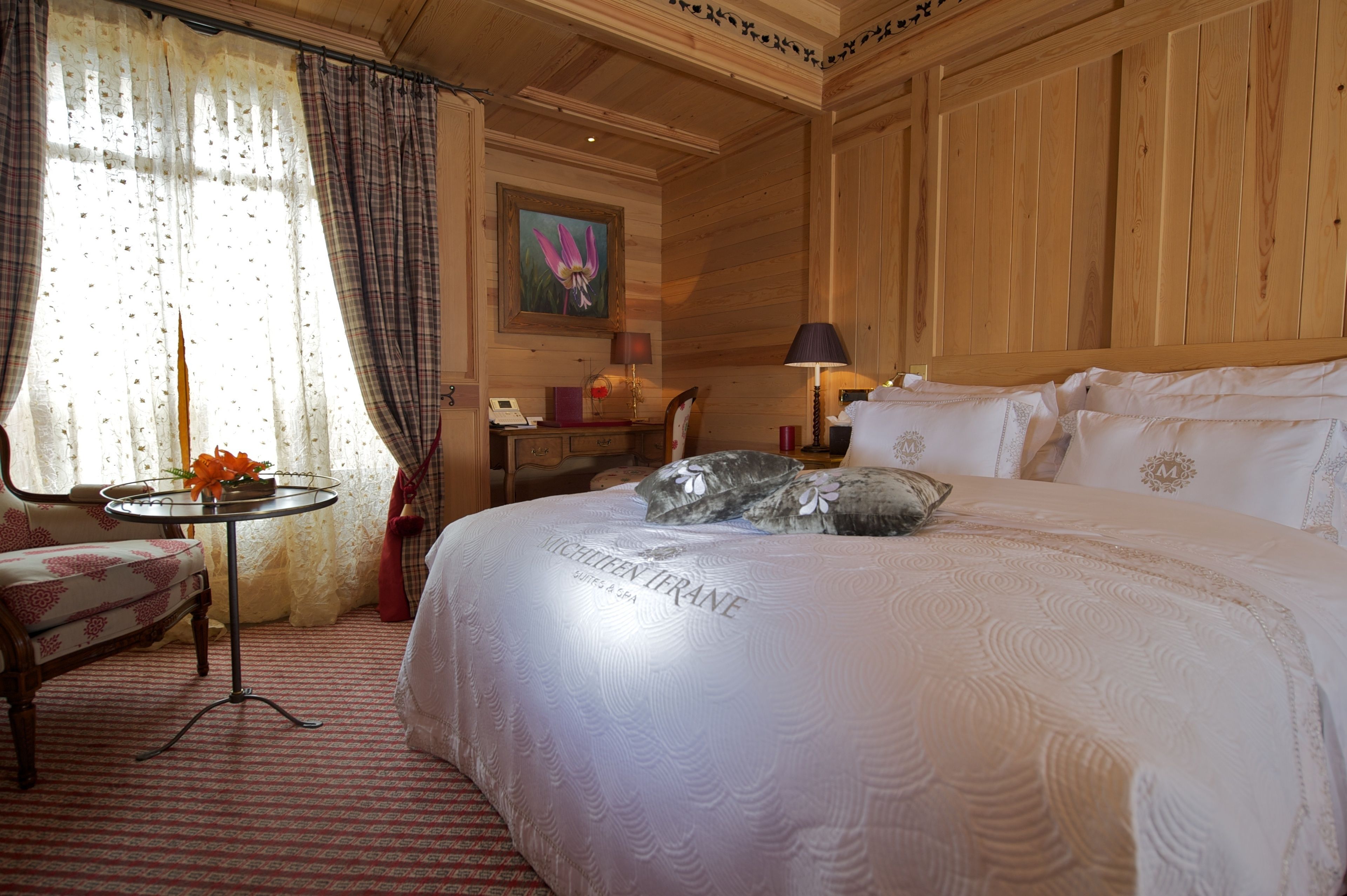 A hotel room with a large bed, a small round table with a vase of flowers, a chair, a desk with a lamp, and a painting on the wall.