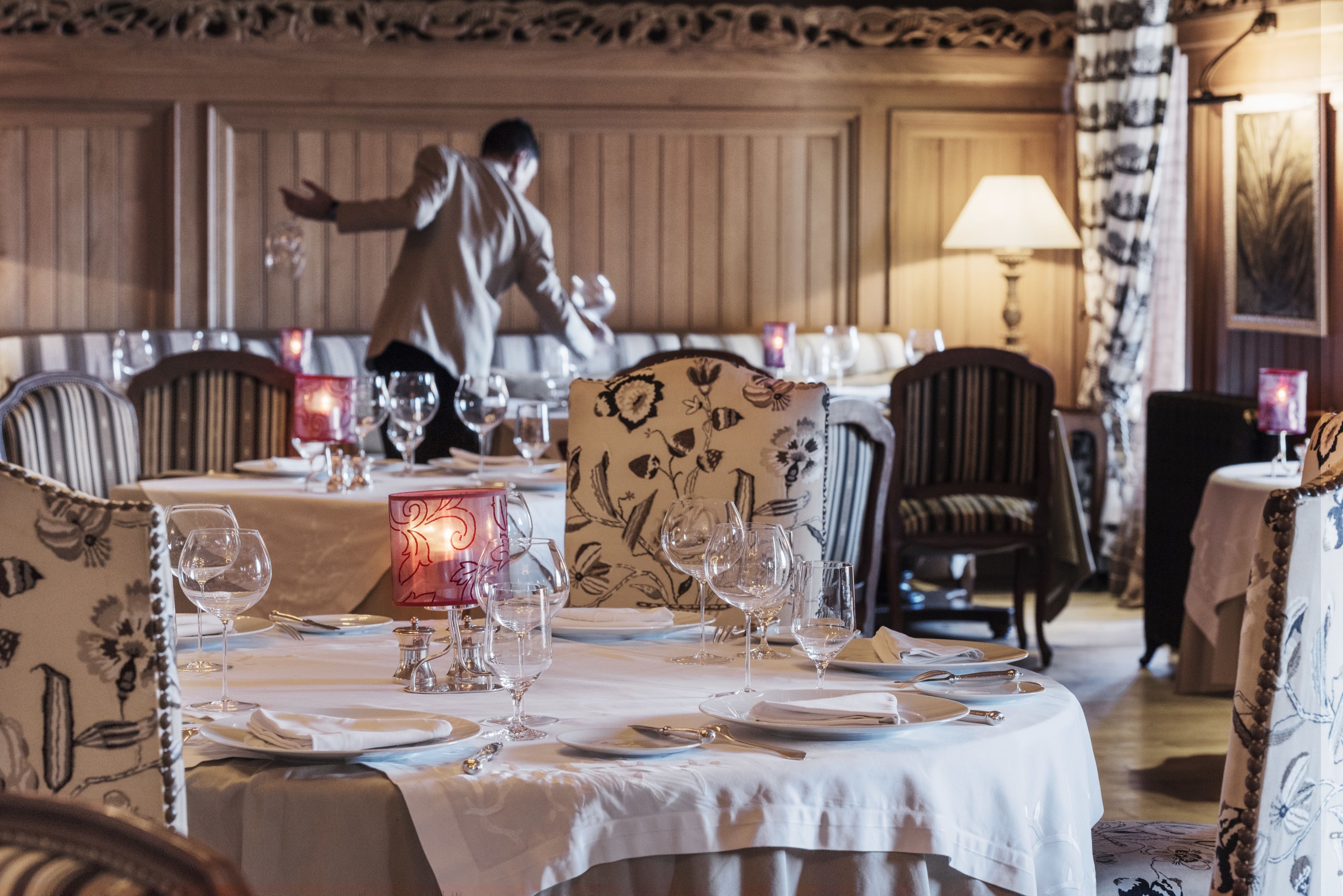 A dining area with tables set for a meal, chairs with floral upholstery, and a person in the background.