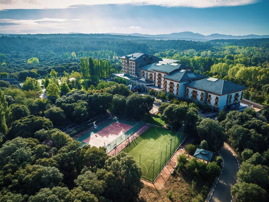 An aerial view of a large building surrounded by dense forest, with a tennis court and a road leading to it.