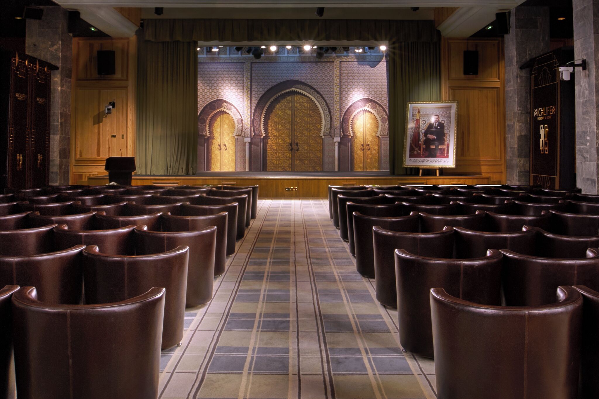 Une salle de conférence avec des rangées de sièges, une scène ornée d’un tableau encadré et des portes richement décorées.