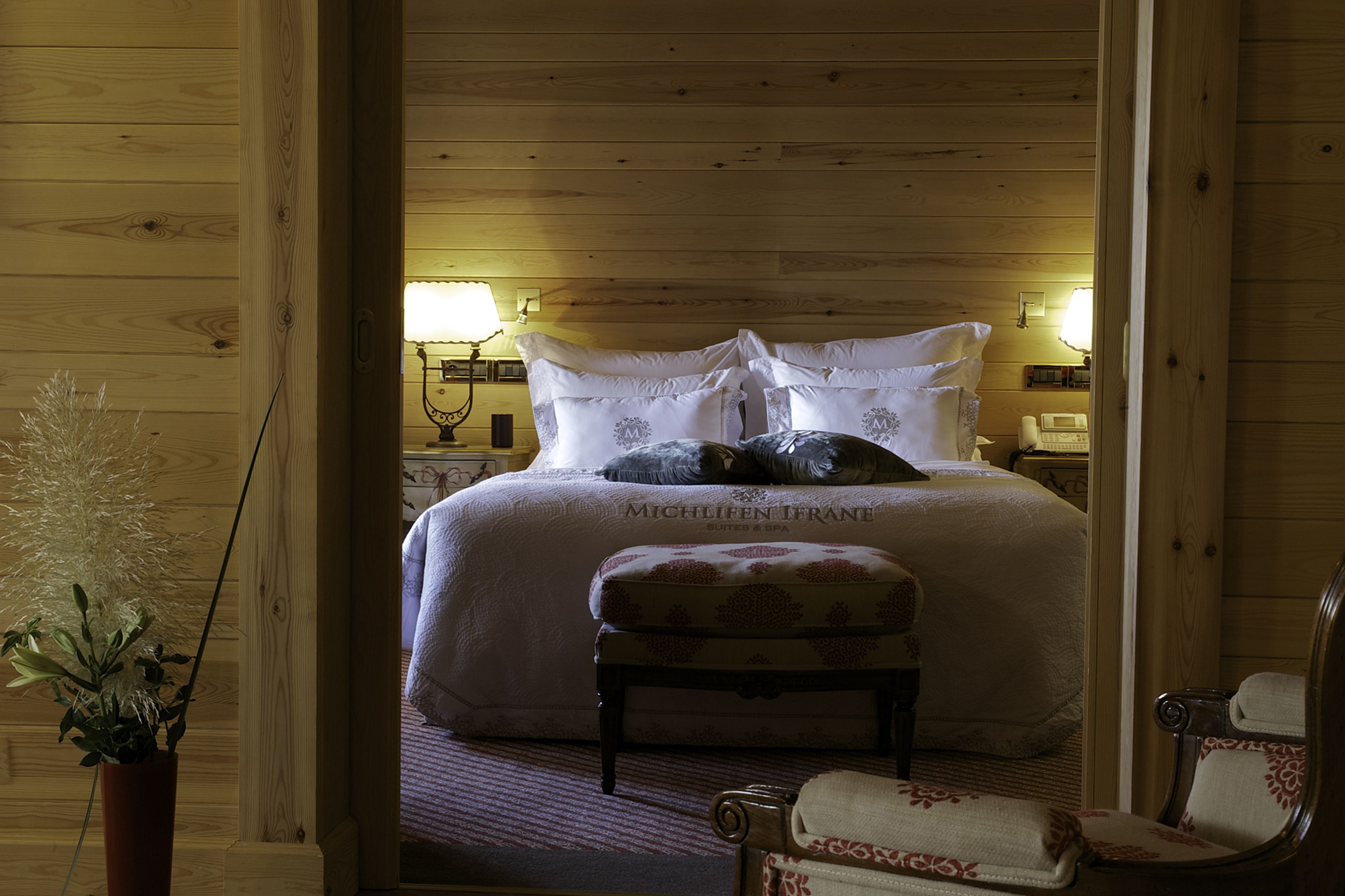 A wooden-paneled room with a bed, bedside lamps, a potted plant, and a chair.