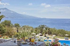 Une table dressée avec de la nourriture et des boissons, donnant sur une piscine et la mer, avec des montagnes en arrière-plan.