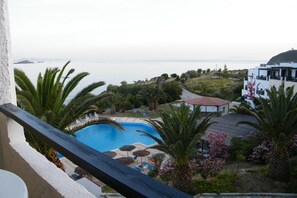 Un espace piscine offrant une vue sur la mer, entouré de palmiers et bordé d’un bâtiment blanc au toit rouge.