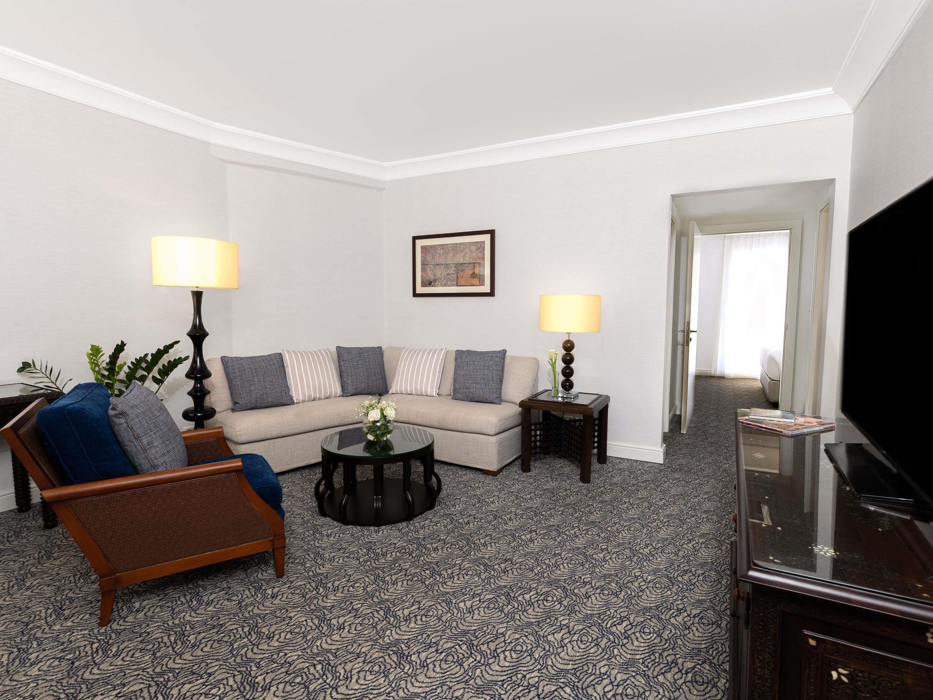 A modern living room with a sofa, armchair, coffee table, and lamps. There is a flat-screen TV and a framed picture on the wall.