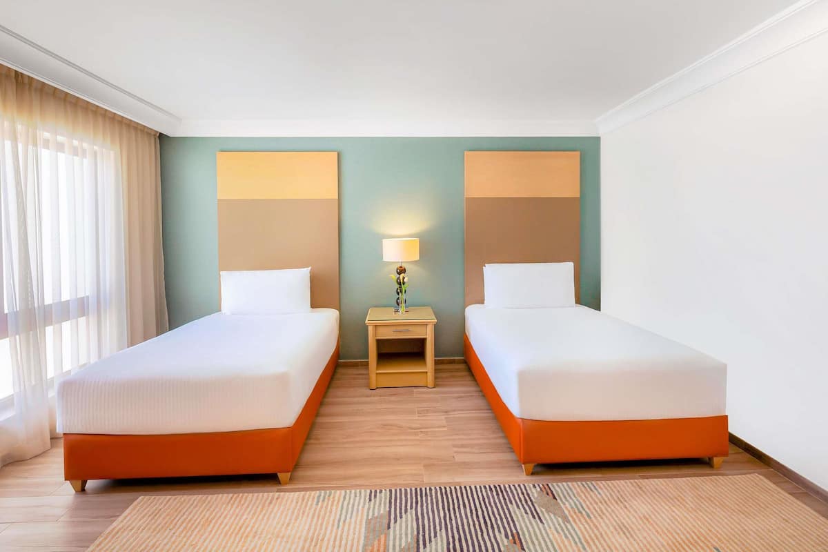 A hotel room with two beds, a nightstand with a lamp, and a window with curtains.