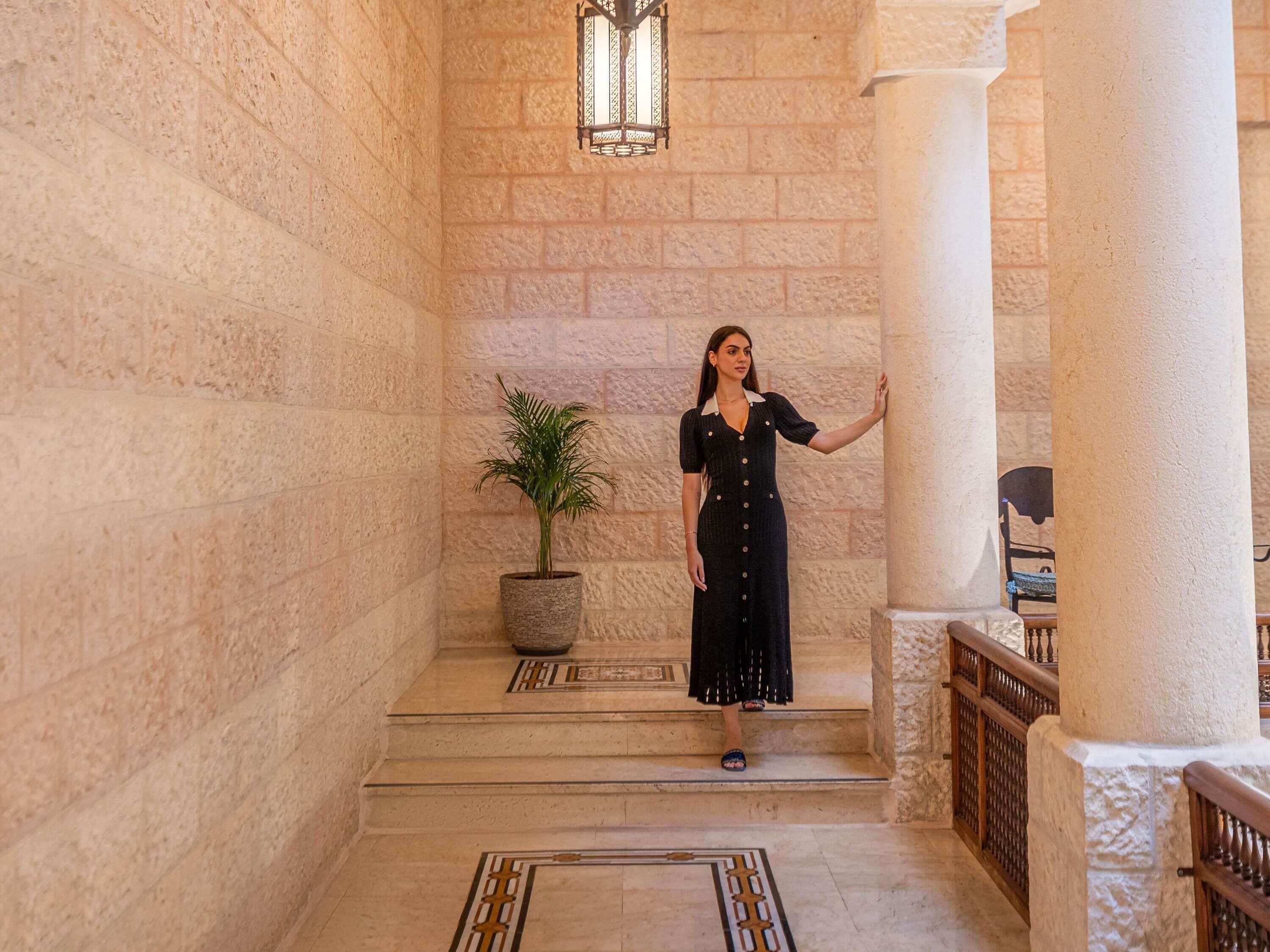 A woman standing on a staircase in a building with stone walls and a potted plant.