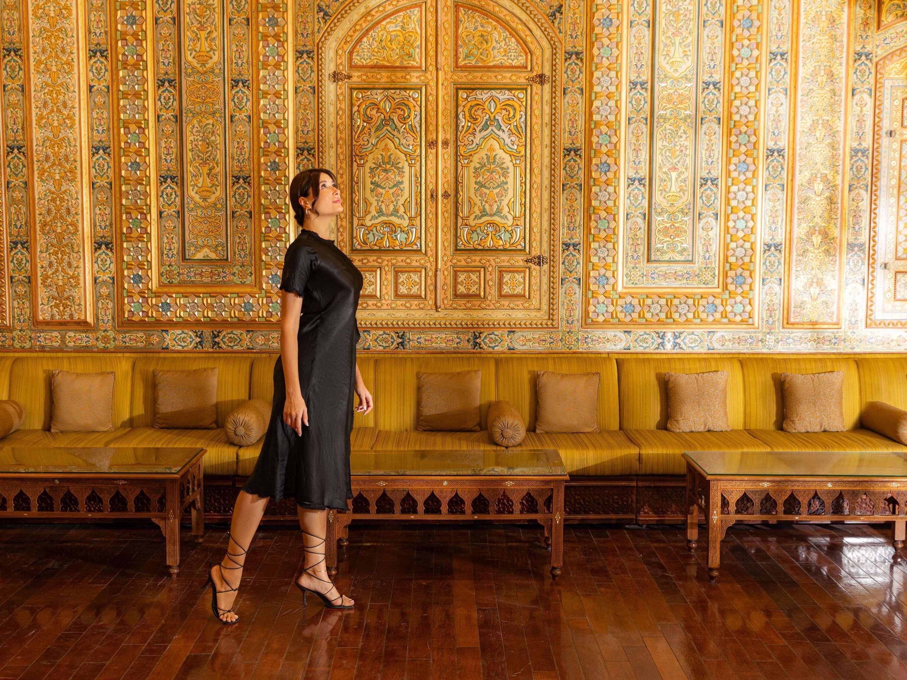 A woman in a black dress and strappy heels walking in a room with ornate, patterned walls and wooden benches.