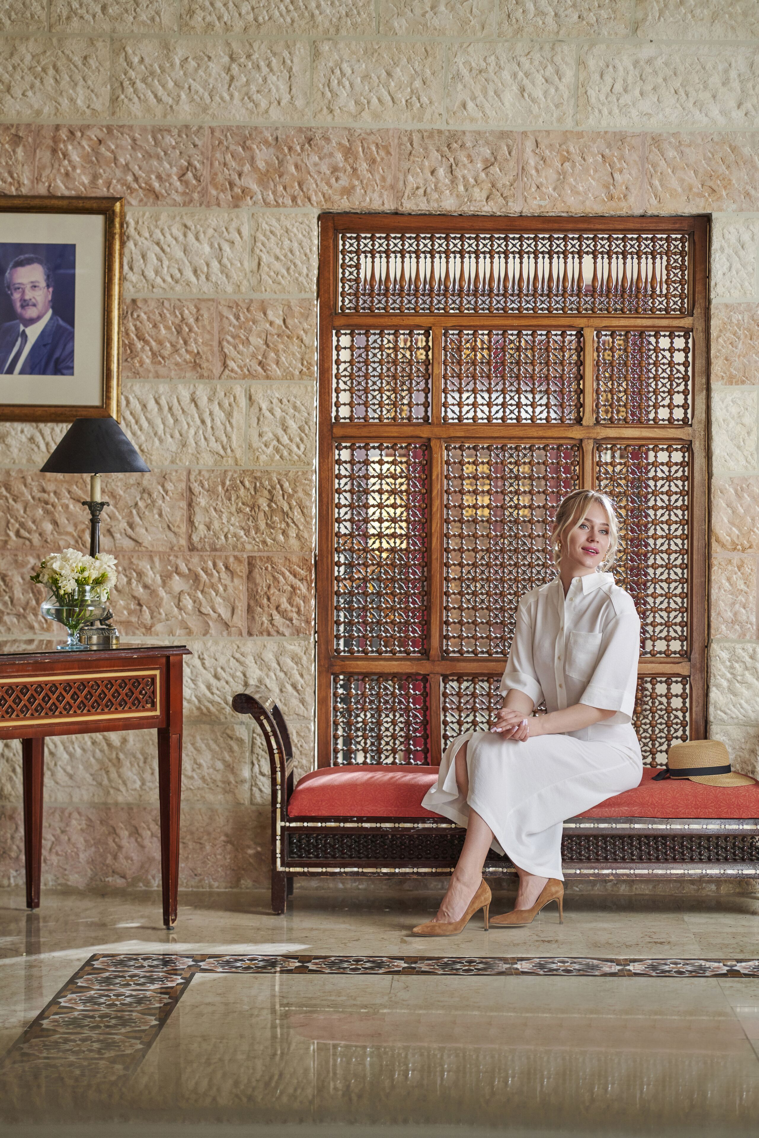 A woman seated on a red cushioned bench in a room with a textured stone wall and a wooden lattice partition. A framed picture hangs on the wall, and a table with a lamp and vase is visible.