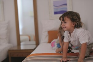 A child crawling on a bed with striped bedding, a white pillow with a pink butterfly, and a wooden nightstand with a lamp.