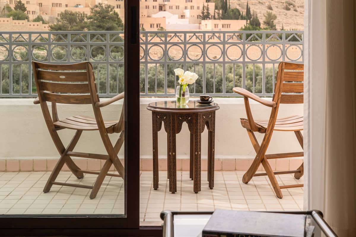 A balcony with two wooden chairs, a small table with a vase of flowers, and a view of a hillside with buildings.