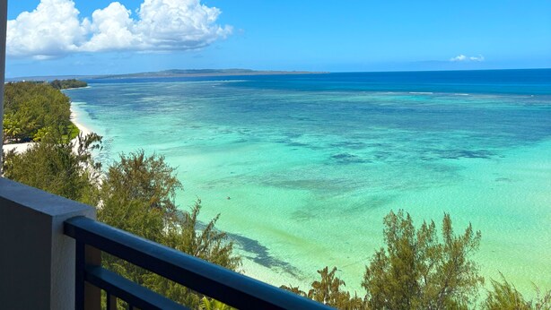 A coastal view with clear turquoise waters, a sandy beach, and lush greenery. - Aquarius Beach Tower (Saipan)