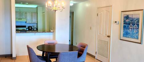 A dining area with a round table and four chairs, a chandelier, a kitchen in the background, and a door with a framed picture on the wall.